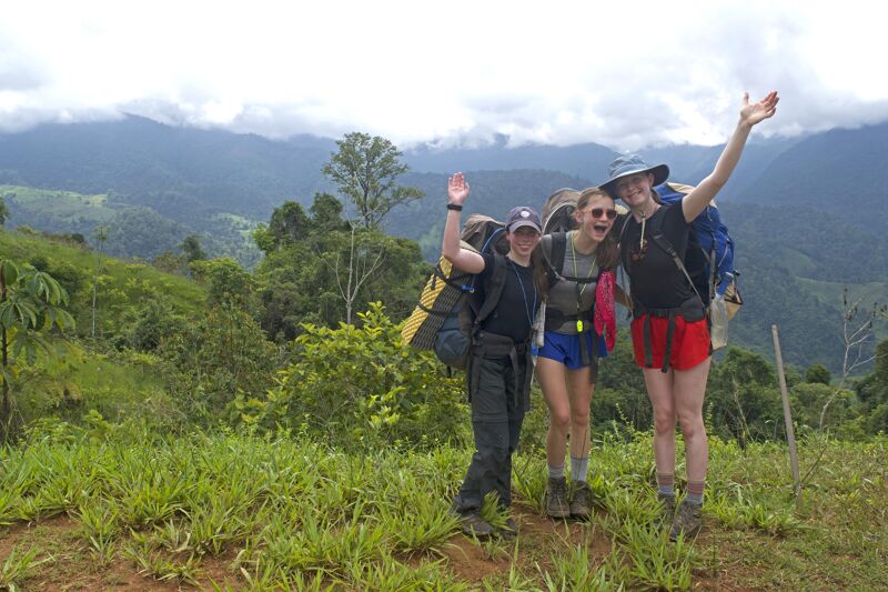 Three young hikers, likely students, are seen on a grassy hilltop, possibly during a break. They are wearing backpacks and hiking attire. The background features a lush, green landscape with rolling hills and mountains under a cloudy sky. The hikers are smiling and waving, suggesting a cheerful and adventurous mood.